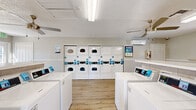 Located at 2848 Caribbean Isle Blvd in Melbourne, Florida, this community laundry facility is a well-appointed, single-floor space designed for convenience and comfort. The room is outfitted with a generous number of top-load washing machines arranged in rows, complemented by a bank of stacked dryer units lining the back wall — all modern, white appliances equipped with digital card-reader payment systems for ease of use.

The interior features light wood-look flooring that lends a clean, contemporary feel throughout. Neutral gray walls with white wainscoting trim add a polished, residential aesthetic. Ceiling fans with integrated lighting provide comfortable air circulation, and a wall-mounted mini-split HVAC unit ensures climate control year-round.

Natural light filters in through multiple windows and glass-paneled doors, keeping the space bright and welcoming. A dedicated waiting area offers a wooden bench and a built-in folding counter, providing ample space for sorting and folding laundry. Additional open shelving is available along the walls for added convenience.

The facility is well-lit with overhead fluorescent fixtures and includes a textured ceiling, a wall-mounted fire extinguisher, and clearly marked exit signage, reflecting attention to safety. The overall ambiance is tidy, functional, and comfortable — a practical shared amenity thoughtfully designed for everyday use.