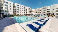 Located at 210 S Main St in Kannapolis, North Carolina, this outdoor amenity space is a well-appointed courtyard nestled within a modern multi-story residential community. The centerpiece of the space is a large, resort-style swimming pool with crystal-clear blue water, flanked by an extensive paver deck lined with teal lounge chairs and in-water sun loungers, ideal for relaxing under open skies. The pool area is enclosed by a sleek black iron fence, providing both security and an elegant aesthetic. Surrounding the pool deck, wide large-format stone pavers extend throughout the courtyard, creating a clean and polished ground surface that ties the space together. Raised planter beds filled with colorful seasonal landscaping, including vibrant red and green foliage, add natural warmth and visual interest to the hardscaped environment. The building facades framing the courtyard feature a contemporary mix of warm taupe horizontal siding and crisp white paneling, with private balconies overlooking the amenity area. Glass-panel entry doors provide seamless access from the courtyard into the building's interior. The overall ambiance is bright, open, and well-maintained, offering a welcoming outdoor retreat for residents.