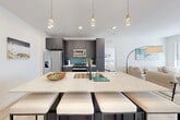 Located at Vintage Commerce in Fort Myers, Florida, this contemporary single-floor unit offers a thoughtfully designed open-concept layout with modern finishes throughout. Light-toned hardwood-style flooring runs continuously across the space, contributing to a cohesive and airy aesthetic.

The kitchen serves as a centerpiece, featuring dark charcoal cabinetry with gold hardware, a striking teal subway tile backsplash, and a full suite of stainless steel appliances. A large waterfall-edge quartz island with an undermount sink and bar seating provides ample workspace and casual dining space, illuminated overhead by a trio of elegant pendant lights.

The living area is bright and welcoming, with multiple windows fitted with horizontal blinds that allow natural light to fill the room. A glass-top coffee table and neutral sectional sofa create a comfortable, modern atmosphere, complemented by access to an outdoor area through a black-framed door.

The bedrooms are serene and well-lit, each featuring large windows, ceiling fans, and light-toned flooring. The primary bedroom includes a private en-suite bathroom and a dedicated walk-in closet, while clean white walls throughout the unit maintain a calm, neutral palette ideal for a variety of interior styles.