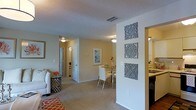 This single-floor residence showcases an open-concept design that connects the living, dining, and kitchen areas into one cohesive and functional space. The living area features plush carpeting in a warm neutral tone, complemented by an orange geometric area rug that adds a vibrant accent. Decorative carved wall panels mounted in a vertical arrangement on a dividing half-wall serve as a standout architectural detail, adding texture and visual interest to the interior.

The dining area accommodates a glass-top table with sleek white chairs, creating an elegant yet casual setting. A framed blue abstract artwork adorns the wall nearby, contributing to the light and airy aesthetic of the space. Flush-mounted ceiling light fixtures provide consistent illumination throughout, while a textured popcorn ceiling adds a traditional finish overhead.

A hallway visible from the main living area leads to additional rooms, suggesting a practical layout designed for easy navigation. The kitchen, partially visible through the open plan, features dark cabinetry that provides a contrasting element to the otherwise light and neutral palette. Overall, the space presents a bright, welcoming ambiance with thoughtful decorative touches.