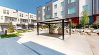 This contemporary residential community showcases a striking modern architectural design, combining corrugated metal panels, smooth stucco, and bold accent colors — including red and orange entry doors — across its multi-story buildings. The exterior aesthetic is clean and urban, with large square windows that allow generous natural light into each unit.

At the heart of the community lies a well-appointed shared outdoor courtyard, designed for both relaxation and social gathering. A sleek, dark-framed pergola with a solid roof panel shelters dual stainless steel grills, creating a dedicated outdoor cooking and entertaining area perfect for residents to gather. The surrounding hardscape features expansive concrete paving that flows seamlessly throughout the courtyard.

Thoughtful landscaping adds warmth and texture to the space, with decorative rock boulders forming natural retaining features, flowering shrubs, ornamental grasses, and young trees softening the urban environment. Pathway lighting bollards line the walkways, providing both safety and ambiance during evening hours.

Individual unit entries are accessible via wide concrete walkways, each with its own address marker and small landscaped buffer. Attached garages provide private parking for residents. The overall community design strikes a balance between modern, low-maintenance living and inviting outdoor shared spaces, fostering a sense of neighborhood under open skies.