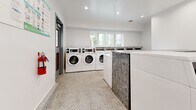 Located in Santa Fe Springs, CA, this shared laundry facility is a compact yet highly functional space designed to serve communal laundry needs. The room is thoughtfully organized to maximize capacity, featuring a row of stacked commercial-grade dryers lining one wall and a series of front-load washing machines positioned along the opposite side, providing ample machine availability for users. The clean white walls and recessed ceiling lighting create a bright, welcoming atmosphere, while the speckled terrazzo flooring adds durability and a polished aesthetic. A window along the rear wall allows natural light to filter into the space, enhancing the open feel despite the room's compact footprint. Two gray entry doors with louvered ventilation panels and automatic closers provide convenient dual-access points, promoting smooth traffic flow. A fire extinguisher is mounted near the entrance for safety compliance, and posted signage offers laundry instructions and best practices for users. The electrical panel is also accessible within the room. Overall, the facility presents a clean, modern, and well-maintained environment equipped with commercial-grade machines to efficiently meet the laundry demands of its users.