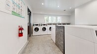Located in Santa Fe Springs, CA, this shared laundry facility is a compact yet highly functional space designed to serve communal laundry needs. The room is thoughtfully organized to maximize capacity, featuring a row of stacked commercial-grade dryers lining one wall and a series of front-load washing machines positioned along the opposite side, providing ample machine availability for users. The clean white walls and recessed ceiling lighting create a bright, welcoming atmosphere, while the speckled terrazzo flooring adds durability and a polished aesthetic. A window along the rear wall allows natural light to filter into the space, enhancing the open feel despite the room's compact footprint. Two gray entry doors with louvered ventilation panels and automatic closers provide convenient dual-access points, promoting smooth traffic flow. A fire extinguisher is mounted near the entrance for safety compliance, and posted signage offers laundry instructions and best practices for users. The electrical panel is also accessible within the room. Overall, the facility presents a clean, modern, and well-maintained environment equipped with commercial-grade machines to efficiently meet the laundry demands of its users.
