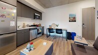 Located at 22 Bay St S in downtown Hamilton, Ontario, this efficiently designed multi-bedroom suite offers a well-organized layout across a single floor. The open-concept living and kitchen area serves as the heart of the unit, featuring exposed concrete ceilings that lend an industrial character, light wood-look flooring, and clean white walls that keep the space feeling bright and airy. The kitchen is equipped with stainless steel appliances — including a full-size refrigerator, stove, dishwasher, and built-in microwave — complemented by sleek dark cabinetry and a light countertop. A dining area with a table and chairs sits adjacent to the kitchen, while the living area provides comfortable seating and a coffee table. Two private bedrooms each offer a bed, a built-in wardrobe with hanging storage, and a dedicated desk and chair setup — well-suited for study and rest. At least one bedroom benefits from a large window with views over the Hamilton cityscape, bringing in natural light and an elevated urban outlook. The full bathroom features a clean, minimalist design with a vanity, toilet, and shower-tub combination. The suite's compact yet functional layout makes it a practical option for student residents near McMaster University.