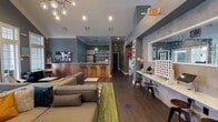This single-floor community clubhouse is thoughtfully designed to serve as a social hub for its residents. The main lounge area is the centerpiece of the space, boasting soaring vaulted ceilings with crisp white crown molding that contrast elegantly against the deep slate-blue walls. Dark hardwood-style flooring runs throughout, grounding the space with warmth and continuity. Large floor-to-ceiling windows with transom panes flood the lounge with natural light, while a set of French doors opens directly to an outdoor pool area, seamlessly blending indoor and outdoor living. The lounge is furnished with generous sectional sofas, accent chairs, and colorful area rugs, creating an inviting atmosphere for relaxation. A wall-mounted flat-screen TV anchors one side of the room. Adjacent to the lounge is a kitchenette and bar area, complete with bar stools, a stainless steel refrigerator, and open shelving. A stylish pendant light cluster adds a modern touch overhead. On the opposite side of the space, a built-in workstation counter set against a textured stone-look accent wall offers individual seating with tablets or devices, ideal for resident use. The counter is accented by a large mirror and decorative elements, including a Connect Four game. The overall ambiance is energetic yet comfortable, blending modern design with playful, community-oriented touches.