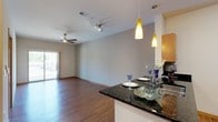 This single-floor unit features a contemporary design with a cohesive, modern aesthetic throughout. The open-concept living area is anchored by rich hardwood-style flooring and neutral gray walls, creating a versatile and welcoming atmosphere. A ceiling fan with integrated lighting overhead and a sliding glass door lead to a private balcony, offering outdoor views and abundant natural light.

The kitchen is seamlessly integrated into the living space via a peninsula counter finished with dark granite countertops, providing both a casual dining surface and a natural division between rooms. Light maple cabinetry lines the galley-style kitchen, complemented by stainless steel appliances including a refrigerator, microwave, and range. Stylish yellow pendant lights above the peninsula add a distinctive design accent.

The bedroom features plush carpet flooring and is served by a generously sized closet with bifold doors and built-in shelving and hanging rods, along with additional storage options provided by surrounding built-in cabinetry.

The bathroom offers a clean, functional design with warm wood vanity cabinetry, white countertops, a large mirror, and a well-lit vanity bar, creating a bright and practical space. Wood-toned doors are consistent throughout, tying the unit's design together cohesively.