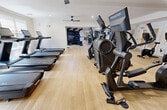 This 1,180-square-foot fitness center is a comprehensively equipped, single-floor facility designed to support a full range of workout routines. The space features light hardwood-style flooring throughout, crisp white walls, and recessed lighting complemented by ceiling fans that promote air circulation during workouts. The layout is thoughtfully organized into distinct workout zones. A dedicated cardio area houses a row of treadmills with individual touchscreen displays positioned along large windows that allow natural light to filter in, offering views of an outdoor pool area. Adjacent to this zone, stationary bikes, stair climbers, and elliptical machines provide additional low-impact cardio options. The strength training section is equally well-appointed, featuring a multi-functional cable machine, a rack of dumbbells ranging across multiple weights, adjustable workout benches, battle ropes, plyo boxes, a wall-mounted rig, and a medicine ball rack. Large mirrors line portions of the walls, enhancing the sense of space and allowing users to monitor their form. A wall-mounted flat-screen television is centrally positioned for entertainment. A built-in water fountain station is conveniently accessible within the facility. The open floor plan maximizes usable space and ensures comfortable movement between equipment zones.