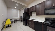 Located at 101 Erie Street in Cheney, WA, Unit 133 is a thoughtfully designed single-floor living space that combines functionality with a clean, contemporary aesthetic. The kitchen is a standout feature, outfitted with rich dark-wood cabinetry extending to the ceiling for ample storage, sleek white countertops, and a full suite of black appliances including a refrigerator, electric range, over-the-range microwave, and dishwasher. Large-format light tile flooring ties the kitchen and adjacent living area together seamlessly. The living area provides a comfortable setup with a gray sofa, coffee table, and a flat-screen TV on a wood-and-metal media stand, all illuminated by natural light from a roller-shade window. The bedroom features soft carpeting, a bed frame with integrated storage drawers, and a dedicated desk and chair — making it well-suited for studying or remote work. A second, similarly appointed bedroom provides additional sleeping space. The bathroom includes a full-size soaker tub and shower combination with clean white surrounds and chrome fixtures. Neutral wall tones and recessed lighting throughout lend the space a bright, modern feel.
