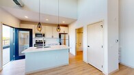This modern loft-style unit showcases an industrial-contemporary design that blends open, airy living with thoughtful finishes throughout. The main living area is defined by impressively tall ceilings with an exposed metallic HVAC duct running overhead, lending an authentic industrial character to the space. Two tiers of black-framed windows — including transom windows above — flood the room with natural light and frame open outdoor views of the surrounding landscape. Light wood-tone flooring extends seamlessly throughout, enhancing the bright, clean aesthetic. The kitchen is well-appointed with crisp white shaker cabinetry, a classic subway tile backsplash, stainless steel appliances including a refrigerator, range, and built-in microwave, and a peninsula with a white quartz countertop and undermount sink. Stylish black cage pendant lights hang above the peninsula, adding a contemporary accent. Adjacent to the kitchen, a full bathroom features a white vanity with storage, a tub-shower combination, and a pendant light above the mirror. A generously sized walk-in closet offers adjustable shelving and multiple clothing rods, providing ample storage. The overall ambiance is bright, modern, and welcoming, with an urban-industrial edge.