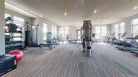 This single-floor fitness center offers a spacious, open layout designed to accommodate a comprehensive workout experience. The interior features neutral-toned walls, recessed lighting, and large ceiling fans that contribute to a clean, airy atmosphere throughout the space.

The facility is generously outfitted with a broad selection of equipment. Along one wall, a row of treadmills, elliptical trainers, and stationary cycling bikes are positioned in front of floor-to-ceiling windows, providing users with bright natural light and scenic views of the surrounding landscape, including palm trees and a water feature. This placement creates a motivating environment for cardio workouts.

The strength training area features a multi-station cable machine, weight benches, free weights, resistance bands, and a variety of selectorized strength machines positioned throughout the open floor. Additional functional fitness accessories, including a stability ball and plyometric box, are also available for dynamic training.

The flooring consists of patterned carpet tiles with subtle striping, adding a polished, professional look to the space. Access to an adjoining room — visible through glass-paneled double doors — suggests additional amenities nearby. The combination of abundant natural light, well-maintained equipment, and a thoughtfully organized layout makes this fitness center a fully functional and welcoming facility.