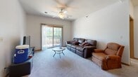 Located at 205 Beach Ave in Ames, Iowa, this single-floor unit offers three bedrooms and two full bathrooms within a well-organized layout. The living room is a comfortable, carpeted space anchored by a ceiling fan and a sliding glass door that opens onto a private balcony overlooking mature trees, allowing natural light to fill the room. The adjacent kitchen features warm honey-toned oak cabinetry, granite-style countertops, black appliances including a refrigerator and electric range, and durable hardwood-style flooring that flows throughout the cooking and dining area. Each of the three bedrooms is carpeted and includes double-door or single closets for storage. The primary bedroom is a well-proportioned space with a double closet and direct access to a full bathroom that includes a bathtub, single-sink vanity with oak cabinetry, and tile flooring. The second full bathroom mirrors this design with a single-sink oak vanity, tile flooring, and clean, neutral finishes. Secondary bedrooms offer windows for natural light and adequate space for standard furnishings. Warm wood-toned trim and doors carry throughout the unit, giving the space a cohesive, understated aesthetic. The overall layout is practical and functional, suited for shared or family living.