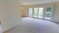 This 2-floor home offers a welcoming and airy atmosphere, highlighted by neutral tones and an abundance of natural light throughout. The main living area is generously sized, finished with soft, neutral-toned wall-to-wall carpeting that adds warmth and comfort underfoot. A classic chandelier serves as a refined focal point, lending a touch of elegance to the open space.

Large French doors dominate one wall of the living area, flooding the room with natural light and providing direct access to an outdoor balcony. The view beyond the doors reveals lush, mature greenery, creating a seamless connection between the interior and the natural surroundings. The light-colored walls and bright windows contribute to a clean, open ambiance throughout the room.

The layout is thoughtfully designed, offering an open flow that makes the living space feel expansive and versatile. The combination of the French doors, chandelier, and soft carpeting creates an inviting environment suited to both relaxed everyday living and entertaining. The two-floor layout of the home suggests additional living or private spaces on the second level, providing a functional separation between common and private areas.