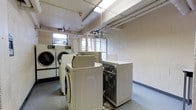 This single-floor laundry facility is a practical, utility-focused space designed to accommodate multiple users simultaneously. The room is lined with painted cinder block walls in a clean white finish, giving the space a straightforward, institutional character. Several top-loading and front-loading washing machines, along with matching dryers, are arranged throughout the room, providing ample laundry capacity. The ceiling features exposed piping and infrastructure, reflecting the functional nature of the space, while a skylight or translucent ceiling panel allows natural light to filter in from above. Additional windows along the walls further brighten the interior, reducing the reliance on artificial lighting. The flooring is a durable, easy-to-clean hard surface well-suited for a high-traffic laundry environment. Visible utility connections and overhead pipe runs underscore the room's operational design. Overall, the space prioritizes practicality and functionality, offering a well-equipped communal laundry area with enough room to move comfortably between machines.