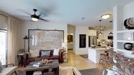 This single-story Flower Mound home showcases an open-concept floor plan that seamlessly connects the living area and kitchen, creating a spacious, cohesive environment ideal for everyday living and entertaining. Light wood-toned flooring runs throughout, lending warmth and continuity to the space.

The living area is anchored by a large-scale framed map artwork that serves as a striking focal point on the main wall. A ceiling fan with a sleek, modern profile provides both function and style overhead. The room accommodates comfortable seating, including a rich brown leather sofa complemented by layered textiles and a rustic wood coffee table, establishing a relaxed yet curated aesthetic.

The kitchen is positioned in an open arrangement with a white peninsula that provides bar-style seating and additional prep space. White cabinetry, pendant lighting, and stainless steel appliances contribute to a clean, contemporary look. Track lighting and recessed fixtures ensure the kitchen remains well-illuminated. A glimpse into an adjacent room reveals additional cabinetry and tiled flooring, suggesting a well-appointed secondary space. The overall ambiance is bright, welcoming, and thoughtfully designed.