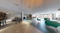 Located at 1 Merrimack Plaza in Lowell, Massachusetts, this single-floor communal living space offers a generously sized, open-plan layout designed for social interaction and relaxation. The space is organized into several distinct functional zones that flow seamlessly into one another, creating a dynamic yet cohesive environment.

The entry area welcomes visitors with glass double doors, pendant lighting, and clean white walls accented by warm-toned flooring. Moving inward, a stylish lounge zone features mid-century modern egg chairs arranged in front of a striking geometric lattice accent wall, complemented by exposed brick detailing and floor-to-ceiling windows that invite natural light and outdoor views.

A media and entertainment wall showcases stacked stone cladding flanked by warm wood shelving, housing flat-screen televisions and decorative objects beneath industrial-style pendant lights. A game area includes a billiards table set against exposed brick, adding a lively recreational dimension.

Separate dining and collaborative seating areas are furnished with long communal tables and industrial-style chairs, positioned near large glass doors that open to outdoor views. Teal-toned accent furniture and mixed-material flooring — combining carpet tiles with polished hard surfaces — unify the space's contemporary aesthetic throughout.