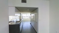 This contemporary two-floor space showcases a refined minimalist aesthetic defined by clean architectural lines, an all-white palette, and high-quality modern finishes throughout. The kitchen is a standout feature, outfitted with stainless steel countertops, a double-basin sink, and full-sized appliances including a refrigerator and range. Sleek, handle-free cabinetry in crisp white extends along the walls, maximizing storage while maintaining the space's streamlined look. Dark tile flooring grounds the interior, providing a striking contrast to the bright white walls and ceilings. A large glass panel partition elegantly separates the kitchen from the adjacent living area, preserving an open, airy feel while defining distinct functional zones. The living area benefits from a generous bank of horizontal windows that span the far wall, inviting abundant natural light and offering views of the surrounding environment. Recessed ceiling lighting supplements the natural light, ensuring the space is well-illuminated throughout. The overall ambiance is one of understated sophistication — a carefully considered design that balances functionality with a clean, modern sensibility across both floors.