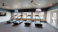 Located on N Pecan Pkwy in Justin, TX, this fitness center is a thoughtfully designed, single-floor facility that offers a full range of workout options in a modern, welcoming environment. The space is divided into distinct functional zones, each catering to different aspects of physical fitness.

The cardio area is prominently featured, housing multiple treadmills and an upright stationary bike positioned along a wall of large windows that overlook an outdoor pool. This layout provides natural light and an engaging view, enhancing the workout experience. Ceiling fans throughout the space ensure comfortable airflow, complementing the recessed lighting overhead.

A dedicated strength training area includes a well-stocked dumbbell rack with a full range of free weights, an adjustable workout bench, and wall-to-wall mirrors that visually expand the space and assist with proper form. A large flat-screen TV is mounted prominently in this zone. Additional cardio equipment, including elliptical trainers and a stair climber, occupies another section of the facility.

The interior design features a two-tone color scheme with deep slate-blue upper walls contrasting against lighter lower walls, giving the space a sleek, contemporary aesthetic. Durable, dark rubberized flooring runs throughout, providing both safety and practicality. Glass-paneled doors connect to adjacent areas, contributing to an open, cohesive layout.
