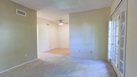 This single-floor residence offers a straightforward and functional layout designed for comfortable everyday living. The interior is characterized by neutral, light beige wall tones that create a calm and cohesive atmosphere throughout. Soft, wall-to-wall carpeting in a complementary neutral shade extends across the living areas, adding warmth and comfort underfoot. The space features an open flow between connected rooms, allowing for a natural transition from one area to the next without visual interruption. A ceiling fan with a light fixture is centrally positioned in one of the main living areas, providing both practical air circulation and overhead lighting. Textured ceilings add a subtle architectural detail, while multiple electrical outlets are conveniently placed along the walls. HVAC vents are integrated into the ceiling, ensuring climate control throughout. The clean, uncluttered layout of the space makes it highly versatile and move-in ready, offering a blank canvas for personalization. Overall, the residence presents a simple, well-maintained interior with a neutral palette suited to a wide range of furnishing styles.