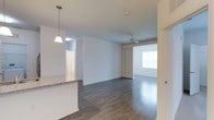 This single-floor space showcases an open-concept layout that seamlessly connects the kitchen and living areas, creating a spacious and airy atmosphere. The flooring throughout is a rich, dark hardwood-style material that adds warmth and a modern aesthetic to the interior.

The kitchen features a raised peninsula countertop finished in speckled granite or stone-look laminate, providing both workspace and casual seating potential. A pendant light with a classic bell-shaped shade hangs above the peninsula, adding a decorative focal point. White cabinetry and bright white walls contribute to a clean, neutral palette that enhances the sense of openness.

The living area is illuminated by natural light streaming through a large window, complemented by a ceiling fan with a light fixture that serves both practical and aesthetic purposes. The layout flows naturally from one zone to the next, with clear sightlines across the space. Smooth white walls and minimal architectural detailing give the interior a contemporary, understated feel. A doorway visible in the background suggests additional rooms, adding to the overall functional versatility of the space.