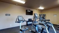 This single-floor fitness room is designed to provide a functional workout environment with a focused selection of equipment. The space features neutral-toned walls and dark carpeted flooring, creating a practical and no-frills atmosphere suited for exercise. Overhead fluorescent lighting ensures the room is well-lit throughout.

The cardio section is prominently featured, housing an upright stationary bike and at least one elliptical trainer, both equipped with digital display consoles for tracking workout metrics. A wall-mounted flat-screen television is positioned centrally, accompanied by a small media console beneath it, allowing users to stay entertained during their workouts. Toward the rear of the room, a multi-use cable strength machine is visible, expanding the workout options available to users beyond cardio alone.

The layout is efficiently organized, making use of the available floor space without feeling overcrowded. The overall ambiance is utilitarian and straightforward, prioritizing functionality. This fitness room serves as a convenient, self-contained exercise facility with essential equipment to support both cardiovascular and strength training routines.