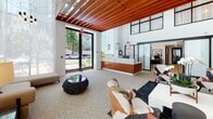 Located at 22121 Clarendon Street in Woodland Hills, Los Angeles, this leasing office presents a refined, design-forward environment that makes an immediate impression. The space is defined by its soaring double-height ceilings, from which rich, warm wood slat paneling extends dramatically overhead, adding texture and architectural depth throughout. Natural light floods the interior through expansive floor-to-ceiling windows dressed in sheer white drapery, creating an airy, welcoming atmosphere.

At the heart of the space, a striking reception desk clad in warm wood cabinetry and topped with a white marble surface serves as a central focal point. Pendant lighting in geometric and linear forms adds a sculptural quality to the ceiling plane. A bold accent wall finished in dark, veined marble provides a dramatic backdrop for a curated seating area featuring eclectic, artfully selected furnishings.

A secondary lounge area showcases built-in wood shelving, framed artwork, and plush mustard-toned sofas arranged around low black tables, creating an inviting, library-like atmosphere. Terrazzo flooring ties the various zones together, lending a polished, cohesive finish. The overall ambiance is one of contemporary elegance, blending natural materials, thoughtful lighting, and curated décor into a highly professional and welcoming leasing environment.