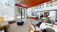 Located at 22121 Clarendon Street in Woodland Hills, Los Angeles, this leasing office presents a refined, design-forward environment that makes an immediate impression. The space is defined by its soaring double-height ceilings, from which rich, warm wood slat paneling extends dramatically overhead, adding texture and architectural depth throughout. Natural light floods the interior through expansive floor-to-ceiling windows dressed in sheer white drapery, creating an airy, welcoming atmosphere.

At the heart of the space, a striking reception desk clad in warm wood cabinetry and topped with a white marble surface serves as a central focal point. Pendant lighting in geometric and linear forms adds a sculptural quality to the ceiling plane. A bold accent wall finished in dark, veined marble provides a dramatic backdrop for a curated seating area featuring eclectic, artfully selected furnishings.

A secondary lounge area showcases built-in wood shelving, framed artwork, and plush mustard-toned sofas arranged around low black tables, creating an inviting, library-like atmosphere. Terrazzo flooring ties the various zones together, lending a polished, cohesive finish. The overall ambiance is one of contemporary elegance, blending natural materials, thoughtful lighting, and curated décor into a highly professional and welcoming leasing environment.