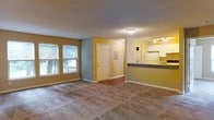 This single-floor residence offers a practical and comfortable layout designed for everyday living. The main living area is spacious and welcoming, finished with plush wall-to-wall carpeting that adds warmth underfoot. Two large windows along one wall invite generous natural light into the room, offering views of the greenery outside. The warm two-tone color scheme — a rich golden yellow on the lower walls and soft gray on the upper portion — gives the space a distinctive and cozy character. A smooth transition leads from the carpeted living area into a hardwood-style entryway, anchored by a classic six-panel front door that provides a clean and defined entry point. Adjacent to the living space, a half-wall pass-through connects seamlessly to the kitchen, which is equipped with white cabinetry, a full-size refrigerator, and track lighting overhead that illuminates the work area effectively. A wall-mounted panel near the entry adds a functional touch to the space. The overall layout is open and efficient, making it easy to move between the living and kitchen areas. The single-floor plan ensures convenience and accessibility throughout, making this a well-appointed and inviting space.