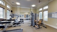 This fitness center offers a well-rounded exercise environment designed to support a variety of workout routines. The space is outfitted with multiple pieces of cardio equipment, including an elliptical trainer and step machines, alongside a multi-station cable weight machine and dedicated strength training benches, providing users with comprehensive options for both cardiovascular and resistance training.

The interior features neutral beige walls accented by crisp white crown molding, lending the space a clean and polished aesthetic. A large mirrored wall runs along one side of the room, visually expanding the space and allowing users to monitor their form during workouts. The flooring consists of a neutral, cushioned carpet suitable for a fitness environment.

Natural light streams in through a bank of large windows fitted with blinds, creating a bright and energizing atmosphere. Overhead, flush-mounted lighting fixtures and a ceiling fan provide additional illumination and airflow, ensuring comfort during exercise. The high ceiling height contributes to an open, airy feel throughout the space. Overall, this fitness center combines practical equipment with thoughtful architectural detailing to create an inviting and functional workout environment.