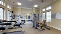 This fitness center offers a well-rounded exercise environment designed to support a variety of workout routines. The space is outfitted with multiple pieces of cardio equipment, including an elliptical trainer and step machines, alongside a multi-station cable weight machine and dedicated strength training benches, providing users with comprehensive options for both cardiovascular and resistance training.

The interior features neutral beige walls accented by crisp white crown molding, lending the space a clean and polished aesthetic. A large mirrored wall runs along one side of the room, visually expanding the space and allowing users to monitor their form during workouts. The flooring consists of a neutral, cushioned carpet suitable for a fitness environment.

Natural light streams in through a bank of large windows fitted with blinds, creating a bright and energizing atmosphere. Overhead, flush-mounted lighting fixtures and a ceiling fan provide additional illumination and airflow, ensuring comfort during exercise. The high ceiling height contributes to an open, airy feel throughout the space. Overall, this fitness center combines practical equipment with thoughtful architectural detailing to create an inviting and functional workout environment.