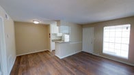 This single-floor space offers a practical and welcoming open-concept layout that seamlessly connects the living area to the kitchen. Rich, dark hardwood-style flooring runs throughout the main living space, providing a warm contrast to the neutral beige walls and white trim. The kitchen is defined by a peninsula counter that serves as a natural divider between the cooking and living areas, offering additional prep space and a casual bar-style seating option. White upper cabinetry and a white refrigerator contribute to a clean, cohesive aesthetic, while the textured ceiling adds subtle architectural character overhead. A flush-mount ceiling light provides functional illumination for the interior. Natural light filters in through a window near the front entry door, brightening the space and enhancing its open feel. The front entry features a solid white paneled door with a multi-point locking system, adding a sense of security and structure to the layout. A fire extinguisher is mounted near the kitchen, reflecting practical safety considerations. Overall, the space presents a straightforward, functional design with durable finishes suited for everyday living.