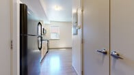 This single-floor unit offers a clean, functional layout designed for comfortable everyday living. The kitchen serves as a central feature of the space, equipped with light maple cabinetry, dark speckled countertops, a double stainless steel sink, and a full suite of black appliances including a refrigerator, dishwasher, electric range, and over-the-range microwave. A window above the counters allows natural light to fill the cooking area. The kitchen flows seamlessly into an open living area, both finished with durable wood-look flooring that extends throughout the unit, lending warmth and visual continuity. The bedroom is bright and airy, featuring a large sliding glass door with sheer curtains that opens to an outdoor area and brings in generous natural light and views of the surrounding exterior. Structural beams add a subtle architectural detail to the ceiling. The bathroom is neatly appointed with a single vanity featuring a dark countertop, a mirrored medicine cabinet, wood-look flooring, and a full tub and shower combination. The unit's neutral, cream-toned walls throughout create a clean and versatile backdrop, making the space feel open, well-lit, and move-in ready.