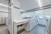 This shared laundry facility is a compact, efficiently organized space spread across a single floor, comprising two connected areas designed for practical, high-traffic use. The interior is finished with large-format white ceramic wall tiles accented by a horizontal metallic border, lending the space a clean, modern aesthetic throughout.

Multiple commercial-grade Huebsch top-load washers and dryers are arranged along the walls, providing ample capacity for shared use. A dedicated folding station with a white laminate countertop and open shelving below sits centrally, offering a convenient workspace for users. A large window running along one wall allows natural light to fill the main area, supplemented by sleek recessed linear LED lighting mounted flush to the ceiling for consistent, bright illumination.

The flooring throughout consists of light-toned porcelain tile in a wood-plank style, contributing to the overall bright and hygienic appearance of the space. Informational signage is posted on the walls, providing clear instructions for machine operation and detergent use. A utility sink, waste receptacles, and a wall-mounted panel are also present, ensuring the facility is fully functional and self-contained. The layout is logical and accessible, with clear circulation pathways between machines, making the space easy to navigate.