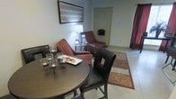 This single-floor space seamlessly blends a dining area with a comfortable sitting lounge, creating a welcoming, multipurpose layout. The dining section features a dark wood round table paired with black leather-upholstered chairs, topped with an elegant candelabra centerpiece and a plate of cookies, lending a warm and inviting atmosphere. Adjacent to the dining area, the sitting space is furnished with two reddish-brown accent chairs accented with nailhead trim detailing, accompanied by a classic white table lamp on a side table. A patterned area rug in warm earth tones defines the seating zone atop the light-colored tile flooring that runs throughout the space. A framed piece of wall art with a structured, linear design adds a decorative focal point above the seating area. The entry door, positioned toward the back of the space, is accompanied by a wall-mounted mailbox, suggesting this is a self-contained unit. A red curtain panel on the right side adds a bold pop of color, contributing to the overall warm and cozy ambiance. The neutral wall tones and thoughtful furnishing arrangement make this a functional yet stylish living space.