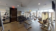 This fitness center is a thoughtfully designed, single-floor facility offering a comprehensive workout environment for users of all fitness levels. The space features rubber flooring throughout, providing both durability and safety underfoot across all exercise zones.

The strength training area is well-stocked, including a multi-station cable machine, a power rack with bench, Raptor-brand equipment, a full dumbbell rack with a variety of weights, and multiple adjustable benches. Functional training is also well-represented, with TRX suspension straps mounted to the wall alongside a collection of stability and medicine balls in various sizes.

The cardio and machine section is positioned near a bank of large windows that flood the space with natural light and offer views to the outdoors, creating an open and airy atmosphere during workouts. Wall-mounted flat-screen televisions are strategically placed throughout the facility, including in both the strength and cardio zones, keeping users entertained and informed.

Full-length mirrors line one wall, enhancing the sense of space and allowing for proper form checks during exercise. Recessed ceiling lighting and pendant-style fixtures provide consistent illumination throughout. The space is accessible via a dedicated entry door and is designed to support a wide range of training modalities in a clean, modern setting.