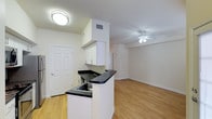 This single-floor space offers a clean, functional layout centered around an open-concept design that seamlessly connects the kitchen and living area. The kitchen features white upper cabinetry, dark countertops, and a peninsula with a built-in sink that serves as a natural divider between the cooking and living spaces. The peninsula's overhang provides an opportunity for casual seating, making it a practical and social focal point. A flush-mount ceiling light illuminates the kitchen area, while the adjacent living space is equipped with a ceiling fan, promoting airflow and comfort throughout. Light hardwood-style flooring runs consistently across the entire space, contributing to a cohesive and airy feel. The white walls and neutral palette enhance the brightness of the interior. A six-panel entry door adds a traditional architectural touch near the kitchen. The open layout maximizes the sense of space, making the unit feel welcoming and versatile for a variety of furniture arrangements and lifestyle needs.