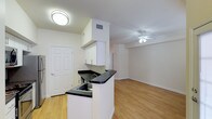 This single-floor space offers a clean, functional layout centered around an open-concept design that seamlessly connects the kitchen and living area. The kitchen features white upper cabinetry, dark countertops, and a peninsula with a built-in sink that serves as a natural divider between the cooking and living spaces. The peninsula's overhang provides an opportunity for casual seating, making it a practical and social focal point. A flush-mount ceiling light illuminates the kitchen area, while the adjacent living space is equipped with a ceiling fan, promoting airflow and comfort throughout. Light hardwood-style flooring runs consistently across the entire space, contributing to a cohesive and airy feel. The white walls and neutral palette enhance the brightness of the interior. A six-panel entry door adds a traditional architectural touch near the kitchen. The open layout maximizes the sense of space, making the unit feel welcoming and versatile for a variety of furniture arrangements and lifestyle needs.