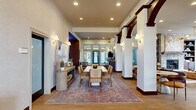 Located at 8495 Bella Woods Drive in Lewis Center, Ohio, this single-floor amenity space offers a generous and well-appointed layout designed to serve as a hub for community gathering and productivity. The living room is a standout centerpiece, anchored by a striking floor-to-ceiling stone fireplace with built-in shelving on either side, complemented by an elegant multi-arm gold chandelier and warm hardwood flooring. Large windows flood the space with natural light and provide views of the surrounding exterior. Adjacent to the living room is a refined dining area featuring a large rectangular table, upholstered seating, and decorative wall art, with direct sightlines to a dedicated theater room marked by its own illuminated signage. The theater room is outfitted with plush recliner seating, wall sconces, and a classic popcorn machine, creating an immersive cinematic experience. The kitchen is clean and functional, featuring white shaker cabinetry with gold hardware, quartz countertops, stainless steel appliances, and hardwood flooring. A beverage and service bar area highlights a dramatic marble-style accent wall with dark wood cabinetry. The office provides a focused work environment with dual workstations and a window overlooking greenery outside.