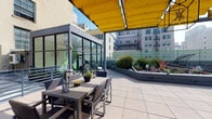 Located at 430 S Broadway in the heart of Downtown Los Angeles, this rooftop terrace offers a compelling urban outdoor experience with sweeping views of the city skyline. The space is defined by its clean, modern design, featuring large-format tile flooring, sleek metal railings, and a striking yellow retractable awning that provides shade over a dedicated outdoor dining area furnished with wicker seating and a wooden table. Lush landscaped planters are positioned throughout the terrace, featuring dramatic agave, cordyline, and ornamental grasses that add texture and color against the urban backdrop. A glass-enclosed structure with floor-to-ceiling dark-framed windows anchors one end of the terrace, housing a fitness area visible from the outdoor space, complete with exercise equipment. The terrace wraps around the building, offering multiple vantage points with unobstructed views of Downtown LA's iconic high-rises and historic architecture. Raised garden beds with varied plantings add a layer of greenery and visual interest. The overall ambiance is contemporary and vibrant, balancing functional amenities with an inviting atmosphere ideal for both relaxation and social gatherings in the heart of the city.