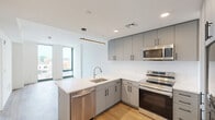 Located on the fourth floor at 801 N Orange St in Wilmington, Delaware, this modern one-bedroom, two-bathroom residence offers a thoughtfully designed open layout with clean contemporary finishes throughout. Light wood plank flooring runs seamlessly through the living areas and bedroom, contributing to a bright and cohesive aesthetic.

The open-concept kitchen and living area serves as the heart of the unit. The kitchen is equipped with sleek grey cabinetry, stainless steel appliances including a refrigerator, range, and microwave, and a large island with a white quartz countertop and undermount sink, illuminated by a modern linear pendant light. The living area benefits from tall floor-to-ceiling windows that flood the space with natural light and offer views of the surrounding Wilmington cityscape.

The bedroom is a well-proportioned retreat, also featuring floor-to-ceiling windows with city views, crisp white walls, and the same warm wood flooring. A hallway lined with open closets on both sides leads to the primary bathroom, which is finished with large-format tile flooring, a wood-toned vanity with a white countertop, and a tub surround clad in dramatic marble-style tile. A second bathroom provides added convenience for guests.