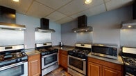 Located at Campus Edge in Carbondale, Illinois, this single-floor communal kitchen is purpose-built to accommodate multiple users simultaneously. The space is defined by its impressive array of stainless steel appliances, including no fewer than four gas ranges, each paired with its own dedicated stainless steel range hood for efficient ventilation. Granite-style countertops run along the perimeter and central cabinetry, providing generous prep space, while warm-toned wood cabinetry below offers ample storage throughout. A full-size stainless steel refrigerator anchors one wall, and a stainless steel dishwasher and double-basin sink are conveniently positioned near a windowed exterior door, allowing natural light to filter in. Additional appliances including microwaves are integrated throughout the workspace. Large windows fitted with vertical blinds along one wall further brighten the kitchen with natural light while offering views of the surrounding neighborhood. The durable slate-style tile flooring adds a practical, easy-to-maintain surface suited for high-traffic use. A drop ceiling with recessed lighting provides even, functional illumination throughout the space. Overall, the kitchen is thoughtfully designed to serve the demands of a shared, community-oriented environment.