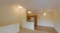 This single-floor space presents an open-concept layout that seamlessly connects the kitchen and living areas, creating a cohesive and functional environment. The kitchen is equipped with warm-toned wood cabinetry that extends to the upper walls, providing ample storage. A white refrigerator and built-in cooktop are integrated into the design, while a peninsula counter extends outward, offering additional workspace and a natural division between the kitchen and the adjacent living area. The flooring in the kitchen transitions to a tile-style surface, contrasting with the light carpeting found throughout the rest of the space. The living area benefits from a ceiling fan with a light fixture, ensuring both comfort and illumination. Recessed and flush-mount ceiling lights provide consistent brightness across both zones. The walls are finished in a clean, neutral tone, contributing to an airy and understated ambiance. The layout is straightforward and practical, with clear sightlines from the living area into the kitchen, making the overall space feel open and connected.