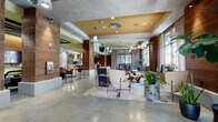 Located at 3111 S Street in Sacramento's vibrant 95816 neighborhood, this contemporary lounge offers a thoughtfully designed, open-plan space that blends industrial and modern aesthetics with warm, inviting accents. Polished concrete floors extend throughout the main area, providing a sleek foundation that contrasts beautifully with rich wood-paneled columns and walls. The ceiling features distinct coffered sections finished in bold gold and green hues, adding visual depth and defining different zones within the open layout.

The main lounge area is furnished with a curated mix of modern seating — including sculptural accent chairs, plush sofas, and organic wood coffee tables — arranged across layered area rugs to create intimate conversation clusters. Large windows along the perimeter flood the space with natural light, while pendant fixtures and recessed lighting provide warm ambient illumination throughout.

A dedicated seating alcove offers a more private retreat, outfitted with ergonomic lounge chairs and teal-toned carpeting beneath a striking green slatted ceiling. Curated artwork adorns the walls throughout, contributing to the lounge's gallery-like character.

The full kitchen is a standout feature, equipped with a large island with bar seating, a stainless steel range hood, high-end appliances, and a vibrant teal ceramic tile backsplash. Cork flooring in the kitchen zone adds warmth and texture. The space is further enhanced by lush indoor plants, contributing to an inviting, layered ambiance.