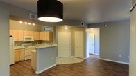 This single-floor residence offers a clean, functional layout with a welcoming open-concept design. The kitchen is equipped with light wood cabinetry, a built-in microwave, a range, and ample counter space along a peninsula that separates the kitchen from the main living area. The peninsula features a light-colored countertop and provides a natural transition between cooking and living spaces. Tile flooring anchors the kitchen zone, while warm wood-look flooring extends throughout the living areas and hallway, adding a sense of warmth and continuity. The neutral gray walls throughout create a calm, versatile backdrop that complements a variety of furnishing styles. A large drum pendant light fixture above the kitchen peninsula serves as a bold focal point. Multiple doorways visible from the main area lead to additional rooms, including what appears to be a hallway corridor accessing further private spaces. Ceiling-mounted light fixtures provide consistent, even illumination throughout. HVAC vents are integrated into the ceiling, ensuring year-round comfort. The overall ambiance is bright, practical, and move-in ready, making it an inviting and straightforward space.