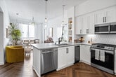 Located in Santa Ana, CA, this well-appointed 3-bedroom, 2-bathroom residence offers a single-level open-concept layout designed for both comfort and style. The kitchen serves as a central hub of the home, featuring crisp white shaker cabinetry with matte black hardware, a geometric tile backsplash, stainless steel appliances including a gas range, microwave, and French door refrigerator, and a large island with bar seating and pendant lighting — ideal for casual dining or entertaining. The kitchen flows seamlessly into the combined living and dining area, where herringbone hardwood floors, abundant natural light, and large windows create a warm, airy atmosphere. The living room is anchored by a sculptural chandelier, while the dining area benefits from a generous picture window with outdoor views. Each of the three bedrooms offers a distinct character — the primary bedroom features a striking textured accent wall and ample closet space, while secondary bedrooms are bright and versatile. The two bathrooms are thoughtfully finished: one includes a glass-enclosed walk-in shower with tile surround, and the other features a tub-shower combo with wood-look tile. Throughout, the home maintains a cohesive, modern aesthetic with clean white walls and warm natural accents.