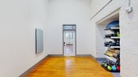 This single-room fitness studio combines historic architectural character with a clean, functional design. The space is defined by its impressively tall ceilings, which create an open, airy atmosphere that feels far larger than its footprint suggests. Warm-toned hardwood floors run throughout, providing a durable and classic surface well-suited for a variety of workouts.

The walls feature exposed brick, painted crisp white, lending the room an industrial yet polished character. A large, multi-pane window along one wall admits abundant natural light and offers views of the surrounding brick building exterior, enhancing the brightness and energy of the space.

A glass-paneled entry door with a transom window above further contributes to the flow of light, maintaining a sense of openness and connectivity. A built-in niche within the exposed brick wall provides organized storage for fitness equipment, including kettlebells, medicine balls, yoga mats, and a stability ball. A wall-mounted mirror panel supports form-checking and movement training, while an additional mounted panel unit serves as an interactive fitness device. The minimalist design keeps the central floor area clear and versatile, making the studio well-suited for personal training, yoga, stretching, or small group fitness sessions.