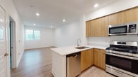 This single-floor space showcases a clean, contemporary design with a cohesive neutral palette and warm natural wood accents throughout. The open-concept layout connects the kitchen directly to the living area, creating a seamless flow ideal for everyday living and entertaining.

The kitchen is a standout feature, equipped with flat-front wood cabinetry extending to the ceiling for generous storage, white quartz countertops, and a full suite of stainless steel appliances including a gas range, microwave, refrigerator, and dishwasher. A peninsula with an undermount sink extends into the space, providing additional prep area and a natural divider between the kitchen and living zone.

The living area is bright and open, benefiting from natural light through a sliding glass door that leads to an outdoor area, complemented by vertical blinds for adjustable privacy. Light hardwood-style flooring runs consistently throughout, enhancing the sense of continuity and space.

A bedroom offers a built-in closet and direct sightlines to the kitchen, maintaining the unit's connected feel. The bathroom is finished with matching wood vanity cabinetry, quartz countertops, a large mirror, and a tub-shower combination with clean white tile surround. Recessed lighting throughout adds a polished, modern touch.