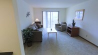 This single-floor space is designed with comfort and functionality in mind. The living area serves as the heart of the home, featuring soft wall-to-wall carpeting that adds warmth underfoot and a neutral color palette throughout that creates a calm, inviting atmosphere. Large sliding glass doors with vertical blinds allow abundant natural light to flood the room while providing direct access to a private balcony, where outdoor seating can be glimpsed beyond. The generous layout of the living area comfortably accommodates a full sofa, armchair, and coffee table arrangement, making it well-suited for both relaxing and entertaining. A built-in shelving unit adds practical storage and display space near the seating area. The clean white walls provide a versatile backdrop that complements a variety of décor styles. Tasteful greenery and accent pieces add life and character to the space without overwhelming its understated design. The overall ambiance is bright, airy, and welcoming, with the seamless connection between the interior living space and the outdoor balcony being a particular highlight, bringing a sense of openness and a pleasant connection to the outdoors.