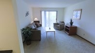 This single-floor space is designed with comfort and functionality in mind. The living area serves as the heart of the home, featuring soft wall-to-wall carpeting that adds warmth underfoot and a neutral color palette throughout that creates a calm, inviting atmosphere. Large sliding glass doors with vertical blinds allow abundant natural light to flood the room while providing direct access to a private balcony, where outdoor seating can be glimpsed beyond. The generous layout of the living area comfortably accommodates a full sofa, armchair, and coffee table arrangement, making it well-suited for both relaxing and entertaining. A built-in shelving unit adds practical storage and display space near the seating area. The clean white walls provide a versatile backdrop that complements a variety of décor styles. Tasteful greenery and accent pieces add life and character to the space without overwhelming its understated design. The overall ambiance is bright, airy, and welcoming, with the seamless connection between the interior living space and the outdoor balcony being a particular highlight, bringing a sense of openness and a pleasant connection to the outdoors.