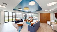 Located at 431 Church St in Norfolk, Virginia, this single-floor communal space offers a well-appointed living and dining area designed with a clean, contemporary aesthetic. Light wood-tone flooring runs throughout, complementing the bright white walls and modern flush-mount circular ceiling fixtures that provide warm, even illumination.

The living room is a welcoming gathering area anchored by a bold blue sofa accented with vibrant throw pillows, a set of cream-upholstered armchairs, and a warm wood side table. A wall-mounted flat-screen TV serves as a central focal point, while a striking tray ceiling painted in deep teal adds a dramatic architectural touch. Potted greenery and curated artwork lend a residential warmth to the space.

The dining area is defined by its dramatic floor-to-ceiling glass curtain wall, flooding the room with natural light and offering expansive views of the surrounding Norfolk cityscape. A sturdy communal dining table paired with terracotta-hued chairs creates an inviting setting for group gatherings.

A glass door opens directly onto an outdoor patio, extending the living space further and providing open-air views of the neighborhood. The seamless connection between interior and exterior, combined with the generous natural light, creates an airy and open ambiance throughout this thoughtfully designed communal space.