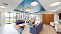 Located at 431 Church St in Norfolk, Virginia, this single-floor communal space offers a well-appointed living and dining area designed with a clean, contemporary aesthetic. Light wood-tone flooring runs throughout, complementing the bright white walls and modern flush-mount circular ceiling fixtures that provide warm, even illumination.

The living room is a welcoming gathering area anchored by a bold blue sofa accented with vibrant throw pillows, a set of cream-upholstered armchairs, and a warm wood side table. A wall-mounted flat-screen TV serves as a central focal point, while a striking tray ceiling painted in deep teal adds a dramatic architectural touch. Potted greenery and curated artwork lend a residential warmth to the space.

The dining area is defined by its dramatic floor-to-ceiling glass curtain wall, flooding the room with natural light and offering expansive views of the surrounding Norfolk cityscape. A sturdy communal dining table paired with terracotta-hued chairs creates an inviting setting for group gatherings.

A glass door opens directly onto an outdoor patio, extending the living space further and providing open-air views of the neighborhood. The seamless connection between interior and exterior, combined with the generous natural light, creates an airy and open ambiance throughout this thoughtfully designed communal space.
